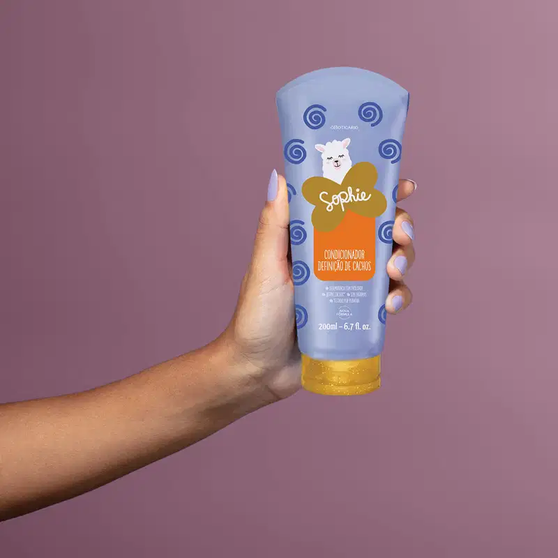 Imagem de um braço com mãos feminina com unhas pintadas de lilás segurando um frasco azul contendo condicionador para cabelos da marca Sophie em fundo roxo.