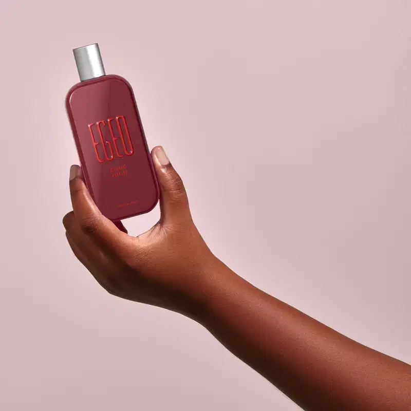 Frasco vermelho de desodorante colônia Egeo Choc High sendo segurado por mão feminina de pele negra contra fundo rosa claro.