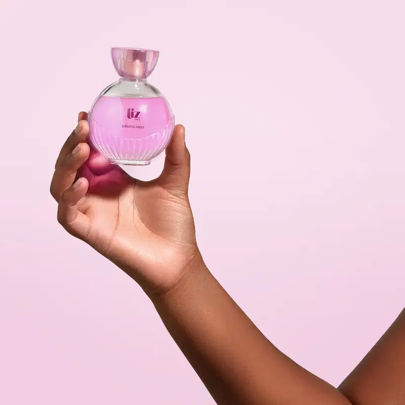 Mão feminina segurando um vidro arredondado com líquido rosa, letras vinho, borrifador dourado e tampa transparente contendo desodorante colônia Liz Flora em fundo lilás claro.