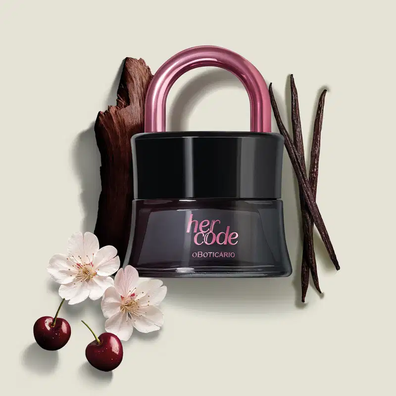 Vidro preto em formato de cadeado com arco e letras na cor rosa metalizado de Eau de Parfum Her Code Touch. Ao redor do frasco, há elementos decorativos como galhos secos, favos de baunilhas, duas cerejas vermelhas e flores de cerejeira rosa claro.