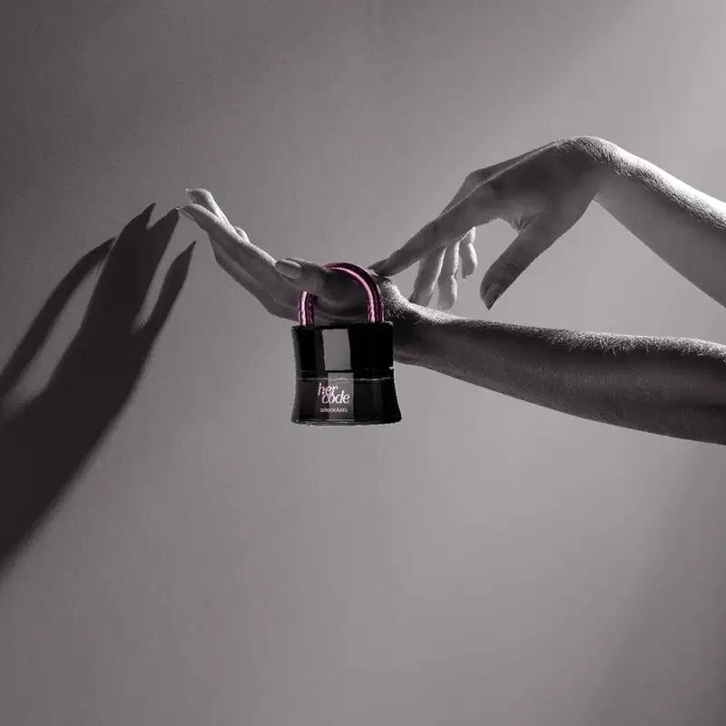Mãos femininas segurando um vidro preto em formato de cadeado com arco e letras na cor rosa metalizado de Eau de Parfum Her Code Touch em fundo cinza escuro com sombras.