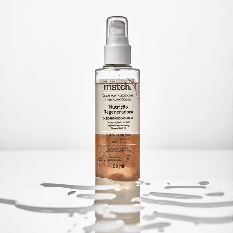 Imagem do um frasco transparente do Óleo Bifásico Capilar Match Nutrição Regeneradora em superfície transparente e molhada. A embalagem possui válvula spray e rótulo laranja com letras pretas. Há respingos de água por todo o frasco.