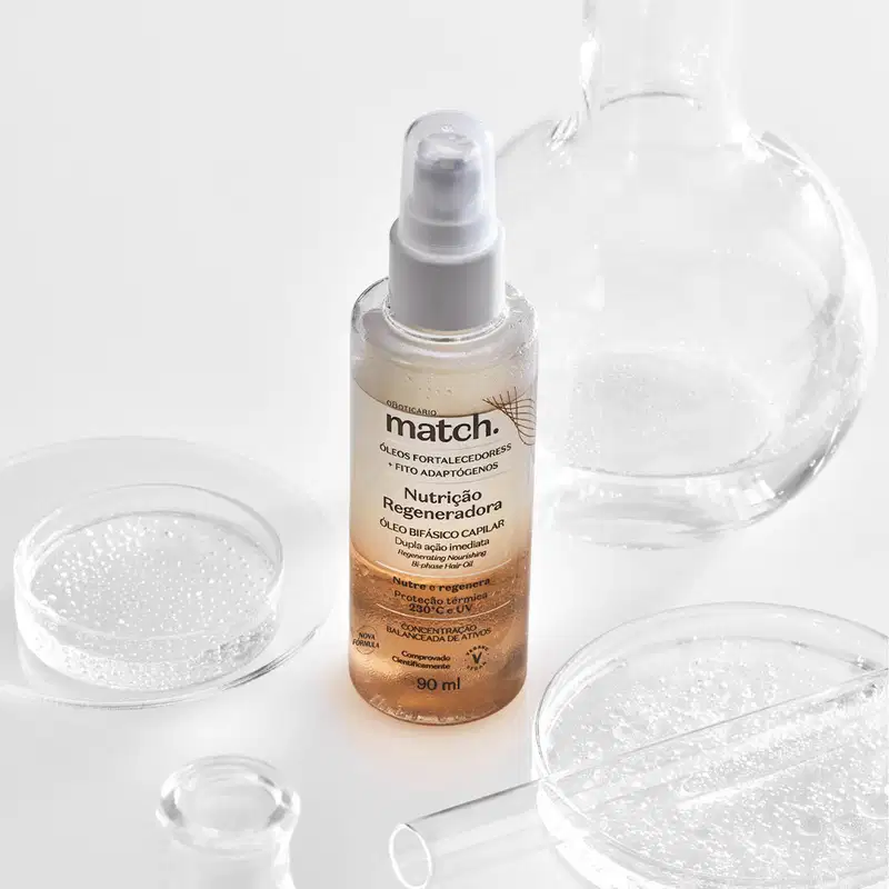 Imagem do um frasco transparente do Óleo Bifásico Capilar Match Nutrição Regeneradora em superfície branca. A embalagem possui válvula spray e rótulo laranja com letras pretas. Ao seu lado estão alguns vidros transparentes.