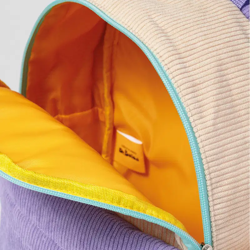 Close-up de mochila infantil de tecido macio em tons de bege e roxo, com zíper azul-turquesa por fora. A mochila está aberta e seu interior é amarelo intenso., 