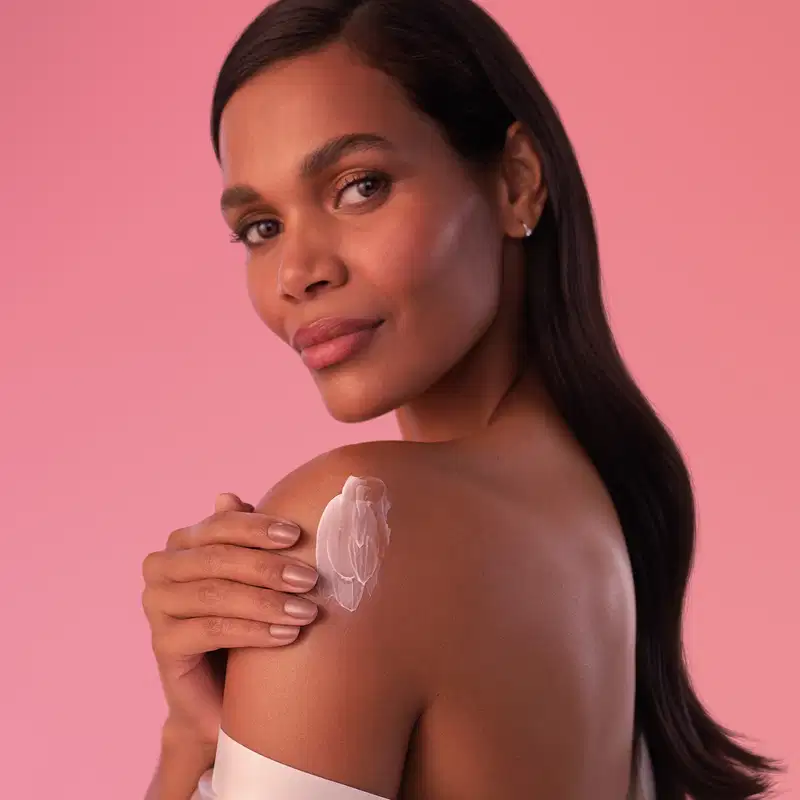 Mulher de pele negra com cabelo longo e liso de perfil passa creme hidratante sobre o ombro em um fundo rosa sólido, com expressão séria e olhar direto para a câmera.