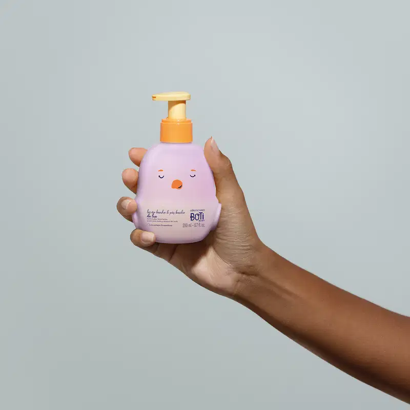 Mão feminina segurando um frasco da loção para banho e pós banho Boti Baby Lua em forma de pintinho na cor lilás e bomba dosadora laranja em fundo lilás.