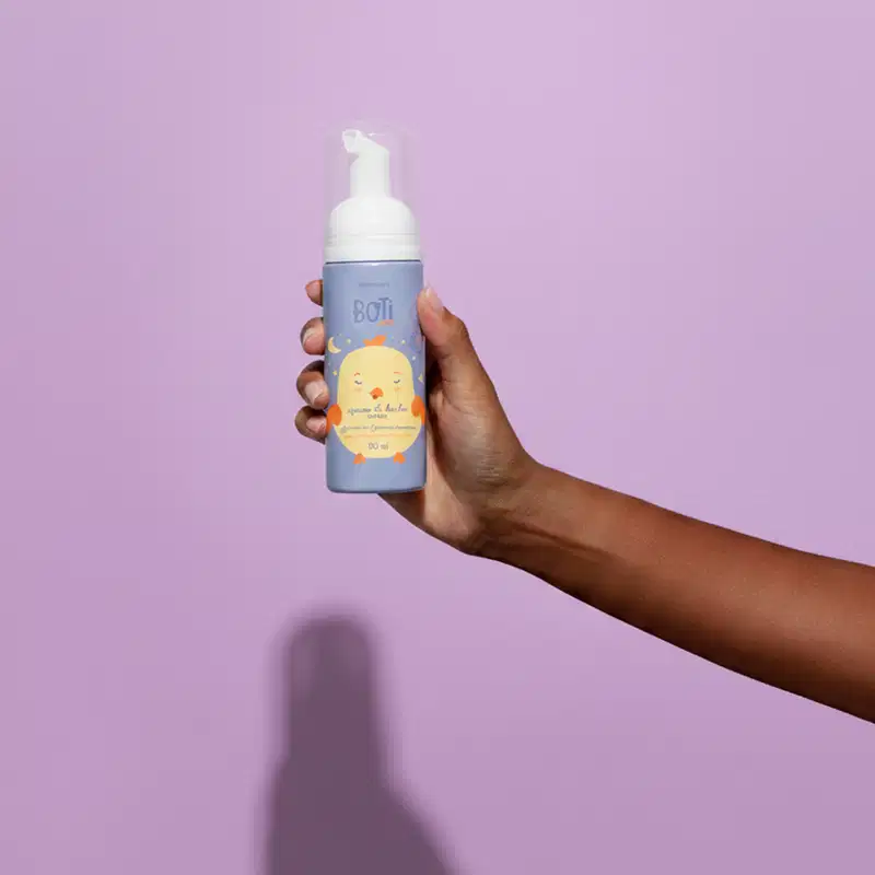 Fundo liso em tons de roxo, com uma mão segurando o frasco da espuma de banho Boti Baby em destaque. A embalagem é azul com tampa branca e possui o desenho de um pintinho amarelo com detalhes em laranja.