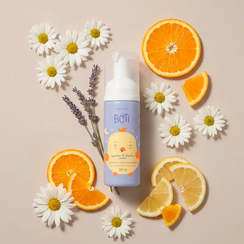 A embalagem azul da espuma de banho Boti Baby é apresentado em um ambiente natural, com flores de camomila brancas e folhas de lavanda, além de fatias de laranja e limão, em um fundo bege claro, sugerindo um contexto de relaxamento e cuidados com a pele.
