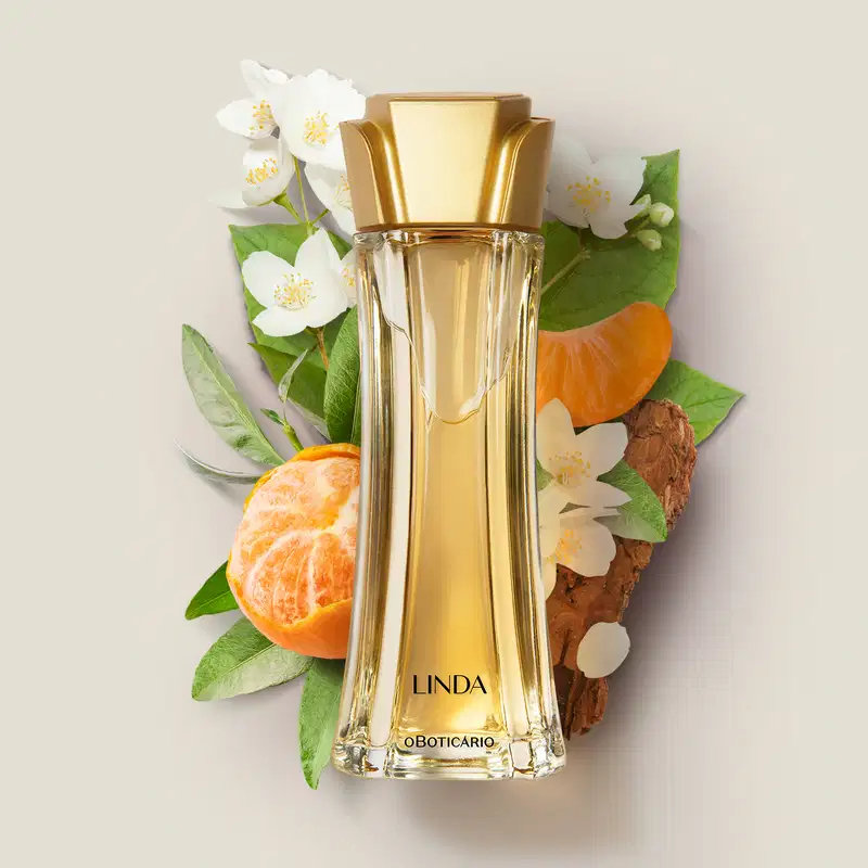 Vidro alongado com líquido bege e tampa dourada em forma de flor de desodorante colônia da marca Linda rodeado por bergamotas, flores brancas e folhas verdes.
