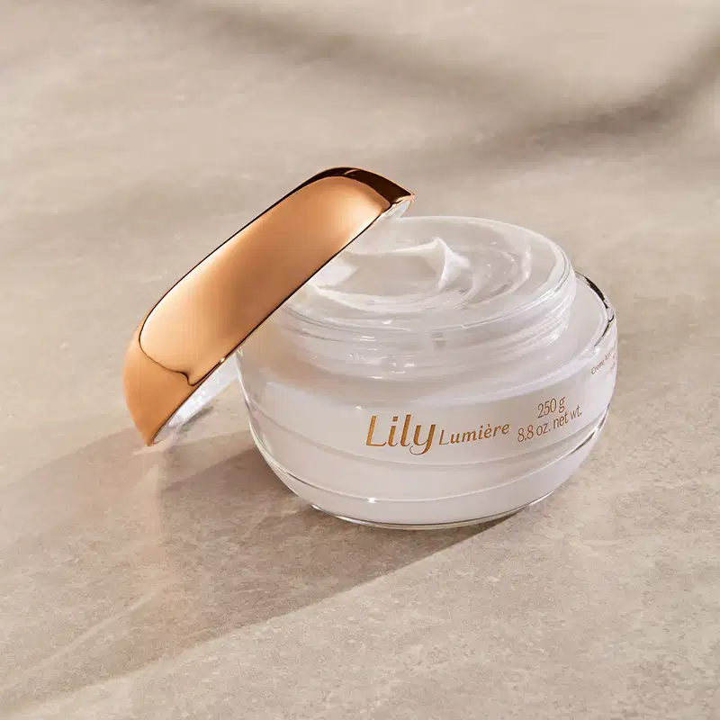 Pote de creme acetinado hidratante corporal Lily Lumière branco com tampa dourada aberto sobre superfície bege clara com sombra, mostrando textura cremosa do produto.