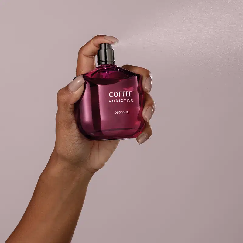 Imagem de uma mão segurando um frasco da marca Coffee Woman Addictive, com a embalagem em tom de roxo escuro e letras brancas", em um ambiente minimalista e monocromático.