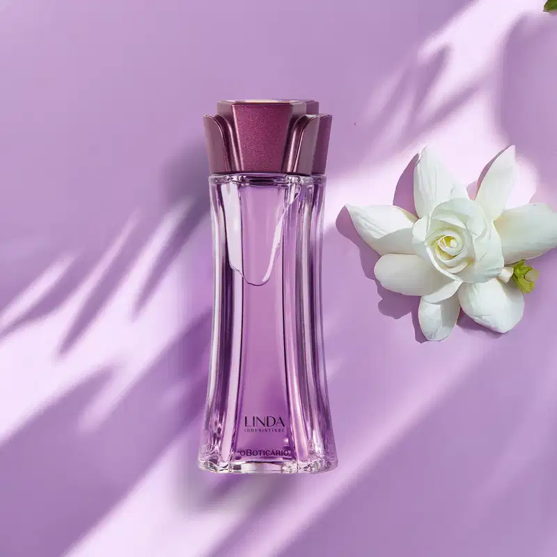 Vidro longo e transparente com líquido lilás, tampa roxa em formato de flor e letras roxas de desodorante colônia Linda Irresistível com camélia branca ao lado em fundo lilás com sombras.