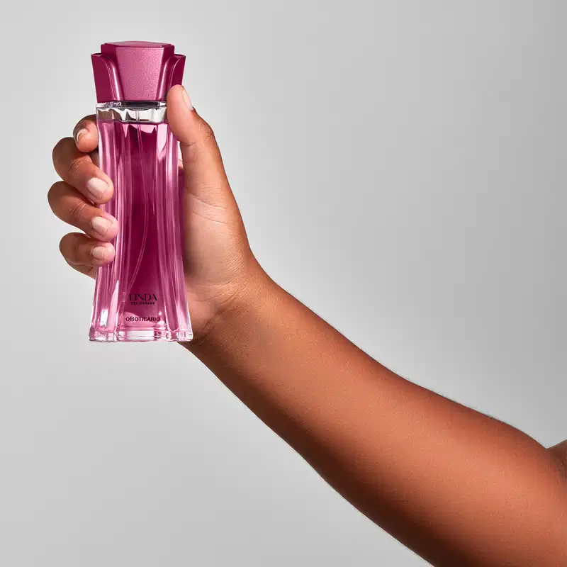 Mão feminina segurando um frasco de vidro com líquido rosa e tampa rosa em formato de flor contendo 100ml de desodorante colônia Linda Felicidade em fundo cinza.
