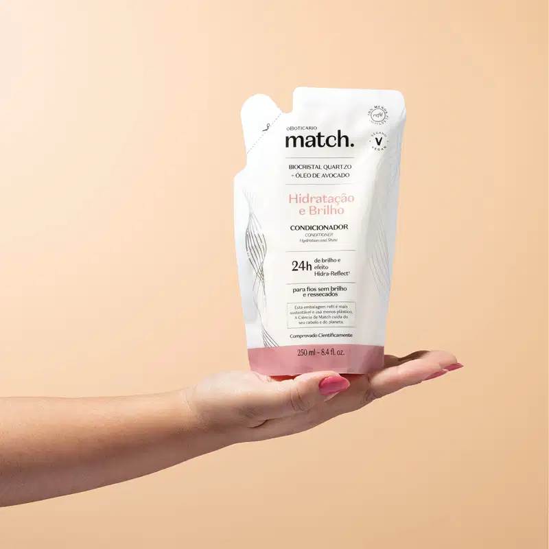 Mão segurando um refil de condicionador Match Hidratação e Brilho em sachê nas cores rosa e branca com letras pretas em fundo em tons de rosa.
