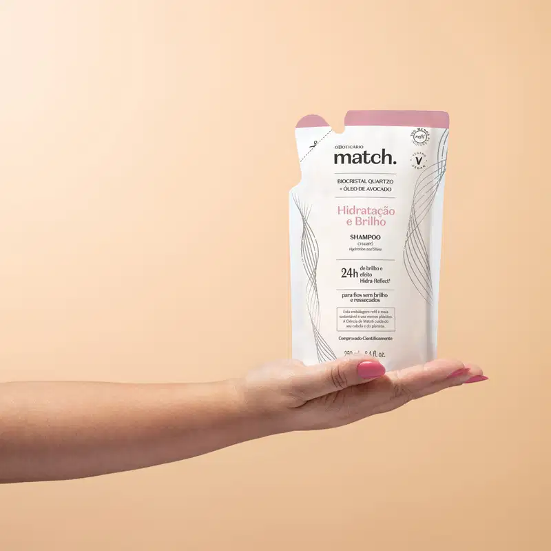 Mão segurando um refil de shampoo Match Hidratação e Brilho em sachê nas cores rosa e branca com letras pretas em fundo em tons de rosa.