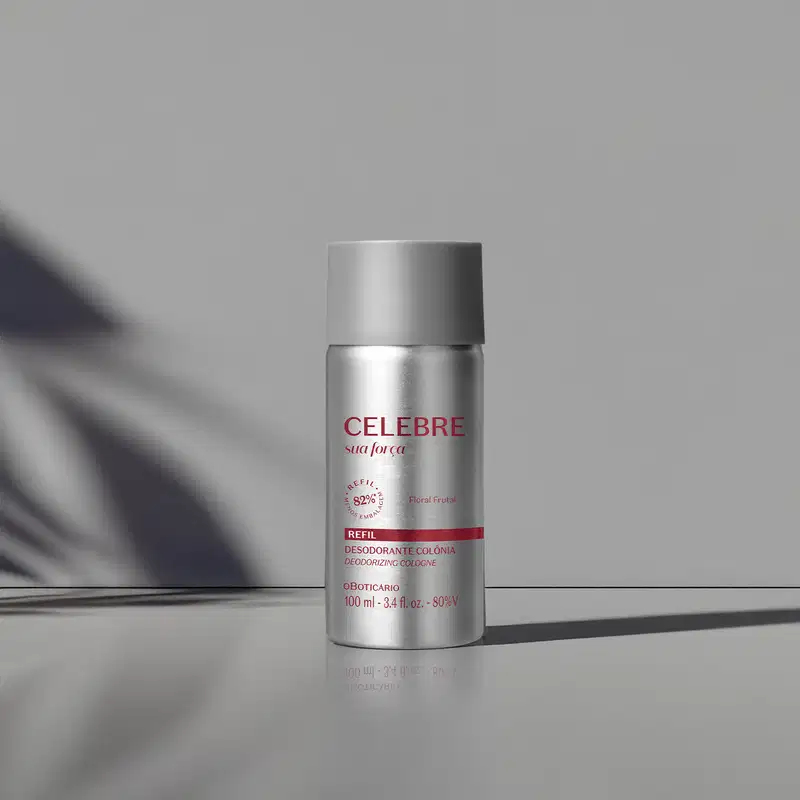 Refil de desodorante colônia Celebre Sua Força Feminino em frasco cilíndrico prateado com tampa cinza e letras vinho sobre superfície cinza com sombra de folha ao fundo, em ambiente com iluminação natural lateral.
