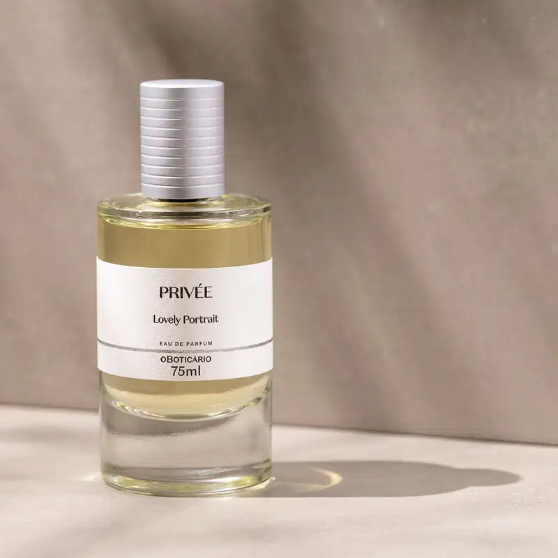 Vidro cilíndrico transparente com líquido bege, tampa prata e rótulo branco com letras pretas de Eau de Parfum Privée Lovely Portrait em superfície clara com sombras e fundo rosado marmorizado.