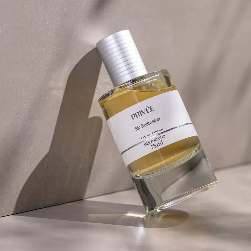 Vidro cilíndrico transparente com líquido alaranjado, tampa prata e rótulo branco com letras pretas de Eau de Parfum Privée Sir Seduction encostrado em um fundo marmorizado bege e superfície da mesma cor.