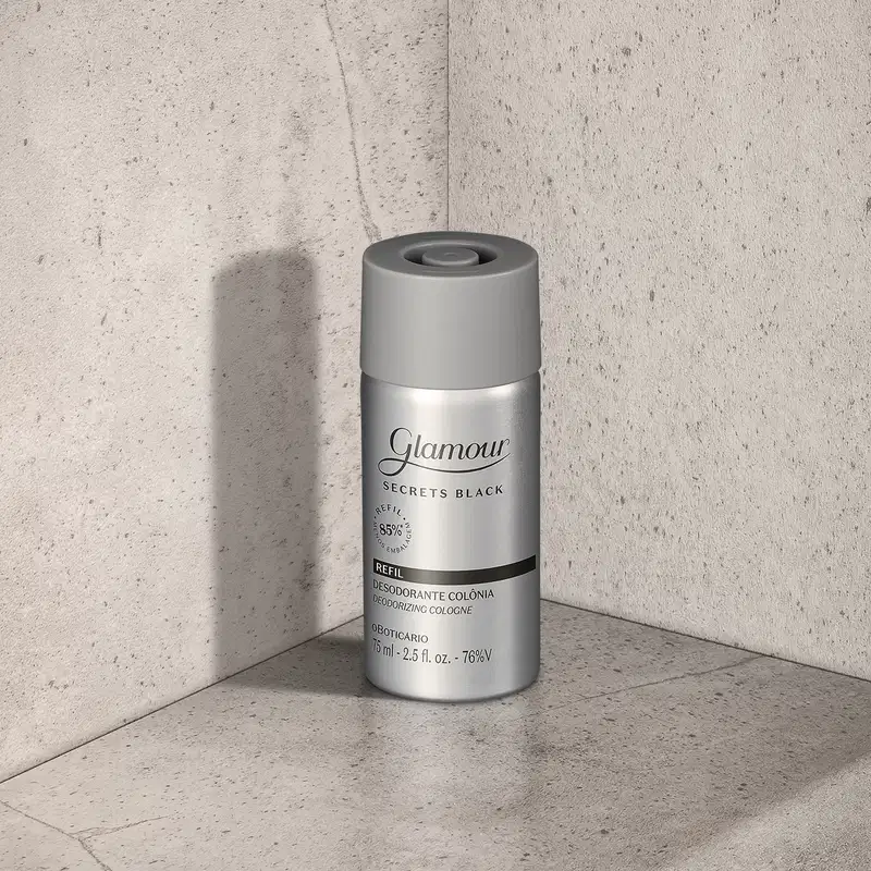 Refil de desodorante colônia Glamour Secret Black em frasco prateado com tampa e letras cinzas e detalhes em preto, posicionado em canto de superfície cinza claro, com parede de concreto ao fundo e sombra projetada à esquerda.