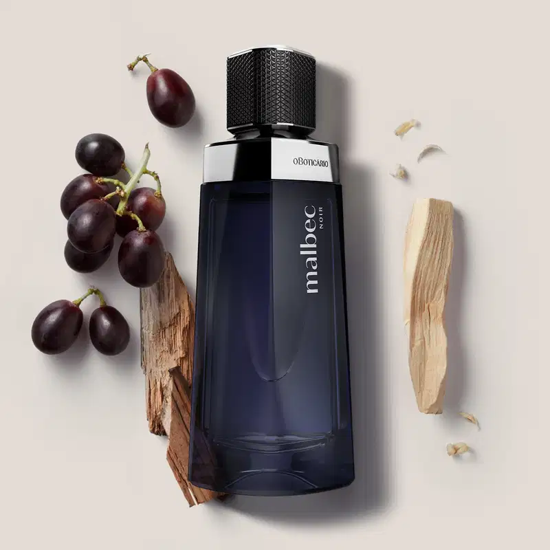O frasco é de vidro escuro com um design geométrico e um tampa preta com detalhes em relevo. Ao lado do frasco, há alguns elementos decorativos, como um pedaço de madeira e alguns cachos de uvas roxas.