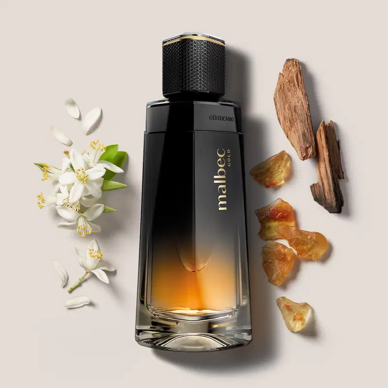 Frasco de desodorante colônia Malbec Gold nas cores degradê de preto com dourado cercado por elementos naturais, como flores brancas e pepitas de âmbar e madeiras, sugerindo ingredientes naturais que compõem a fragrância.