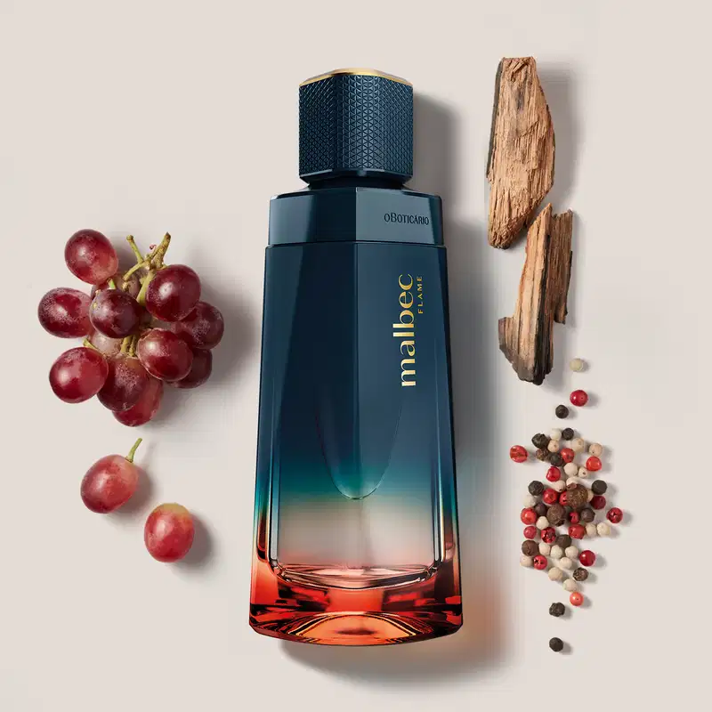O frasco possui um design elegante e moderno, com uma coloração degradê que vai do azul escuro ao laranja. Está posicionado entre alguns ingredientes naturais, como uvas vermelhas e especiarias, sugerindo uma composição aromática.