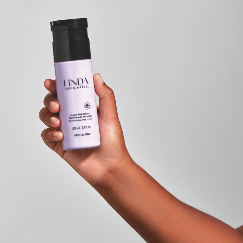 Mão feminina segurando um frasco lilás com tampa e letras pretas de loção hidratante corporal da marca Linda Irresistível em fundo azul claro.