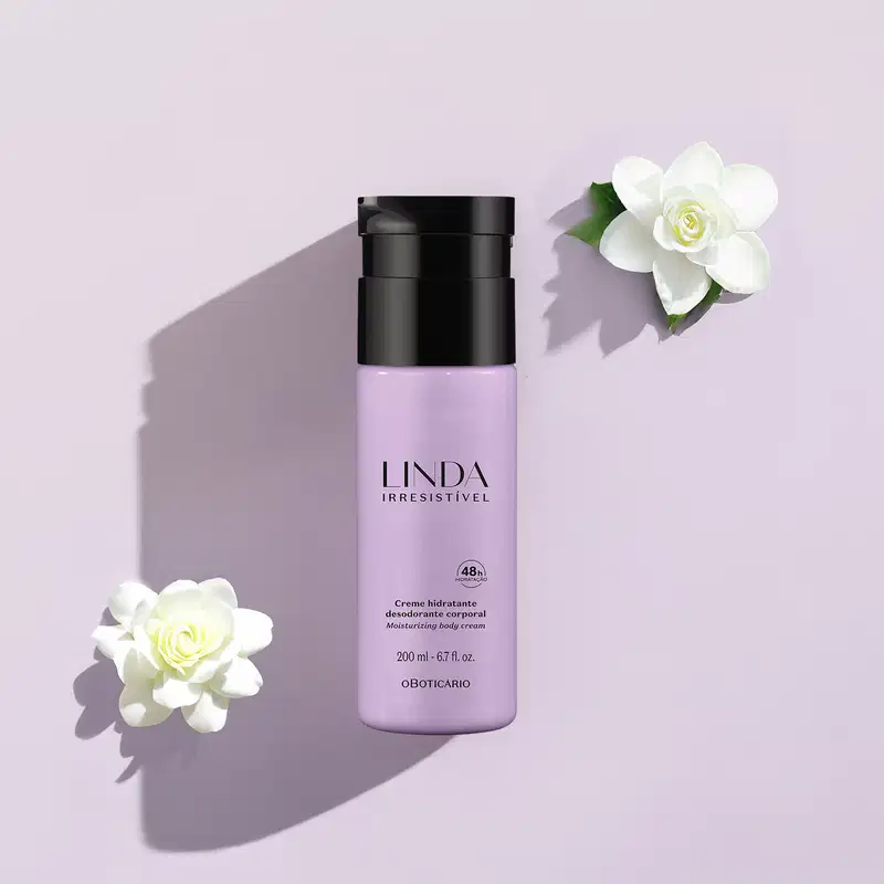 Frasco lilás com tampa e letras pretas de loção hidratante corporal da marca Linda Irresistível com camélias brancas em volta em um fundo azul claro com sombras.