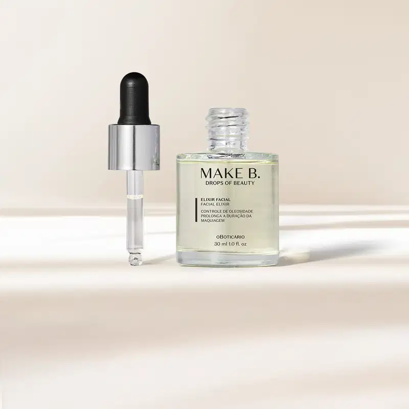 O frasco transparente com líquido claro de Elixir Facial Make B. possui tampa prateada com conta-gotas preto e está em fundo claro com sombras.