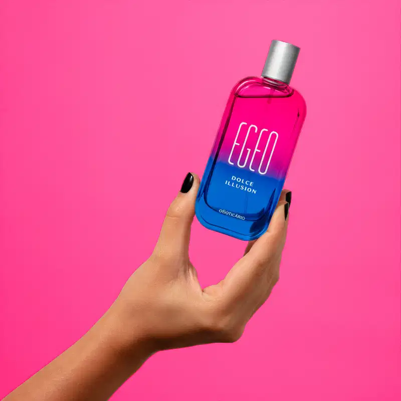Imagem de um recipiente de desodorante colõnia Egeo Dolce Illusion  com design moderno e cores vibrantes de rosa e azul, segurado por uma mão feminina com unhas escuras, em um fundo rosa sólido.