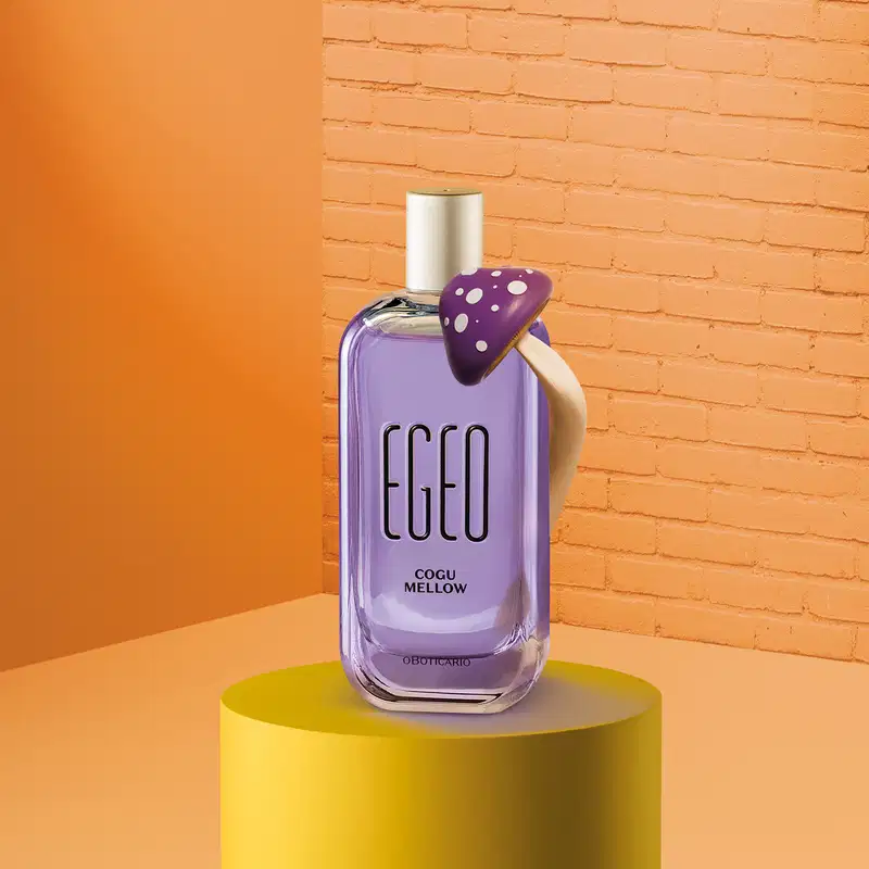 Imagem de um vidro com líquido azul e tampa prata de desodorante colônia Egeo Cogu Mellow, com cogumelo roxo e branco ao lado e fundo em tons de laranja.