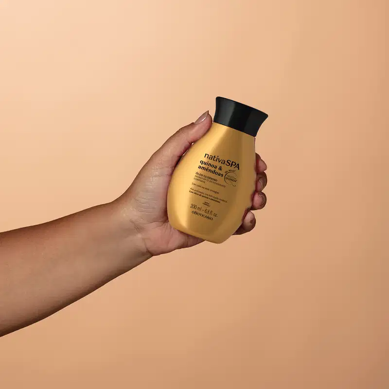 Imagem de um frasco de óleo glorioso corporal da marca Nativa SPA Quinoa & Amêndoas, em tom dourado, sendo segurado por uma mão em fundo laranja-claro.
