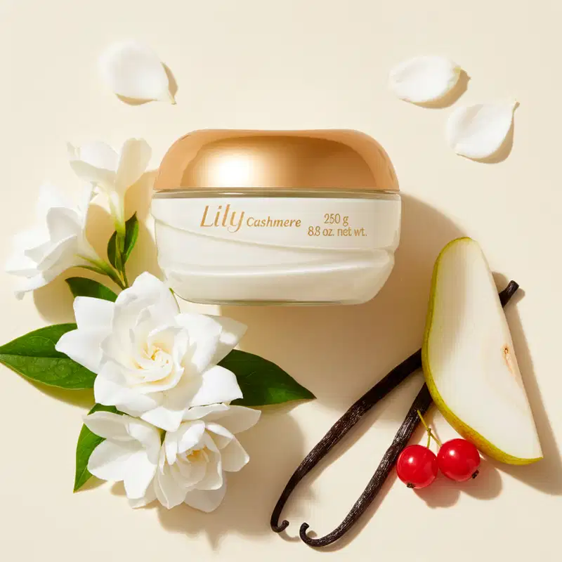 O Creme Hidratante Corporal Lily é apresentado em um ambiente elegante e natural, com flores brancas, frutas e especiarias, sugerindo uma fragrância delicada e sofisticada. A embalagem em tons de branco e dourado destaca a marcado produto.