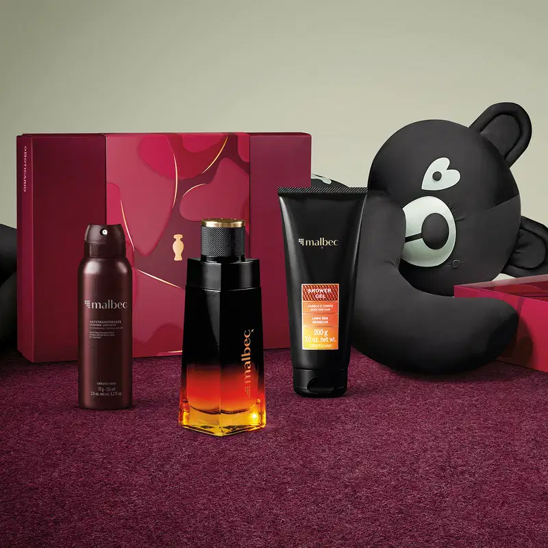 Frasco de desodorante colônia, bisnaga de gel de banho, embalagem de antitranspirante aerossol jato seco e caixa vinho ao lado de um urso de tecido preto e branco em superfície vermelha com floral e fundo cinza.