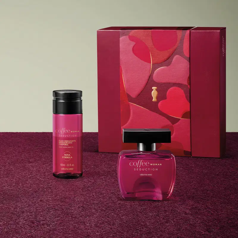Produtos Coffee Woman Seduction, sendo um frasco de desodorante colônia, frasco de óleo hidratante corporal, ambos com líquido rosa e tampa preta, e uma caixa de presente vermelha com detalhes dourados em superfície vermelha com floral e fundo cinza.