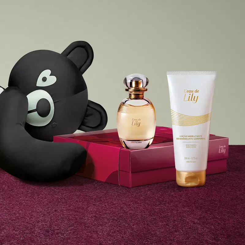 Urso de tecido preto e branco, frasco de desodorante colônia Lily, frasco de loção hidratante corporal da mesma marca e caixa na cor vinho sobre superfície vermelha, em ambiente minimalista com tons neutros.
