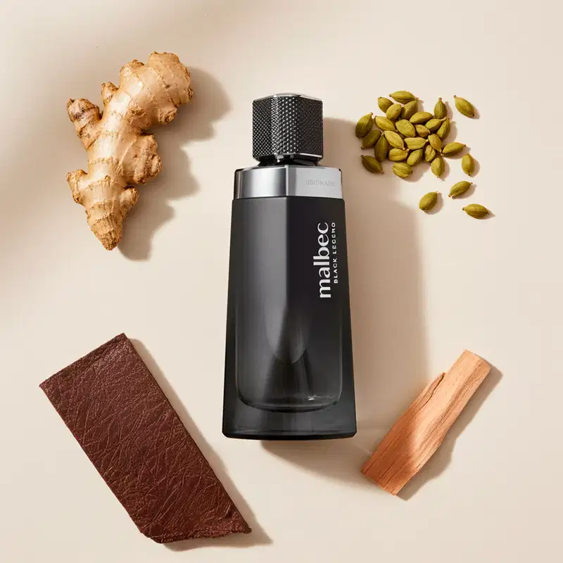 Imagem de uma superfície bege com um frasco preto de desodorante colônia Malbec Black Legend, acompanhado de ingredientes naturais como gengibre, pistache e madeira. O cenário sugere um contexto de cuidados pessoais e bem-estar.