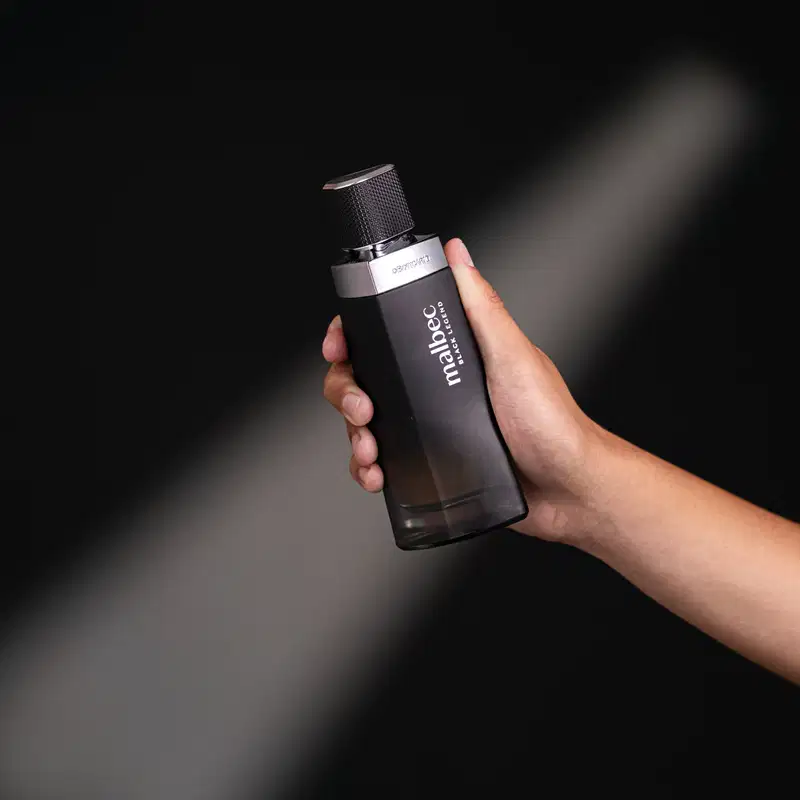 Imagem de um frasco preto de desodorante colônia Malbec Black Legend em uma mão contra um fundo escuro. O produto possui um design minimalista e elegante.
