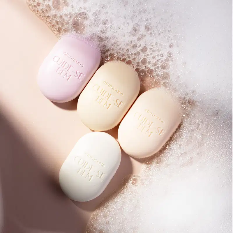 Imagem de quatro sabonetes sortidos da marca Cuide-se Bem em tons de lilás e rosa sobre superfície rosa com espuma branca, sugerindo um ambiente de cuidado pessoal e bem-estar.
