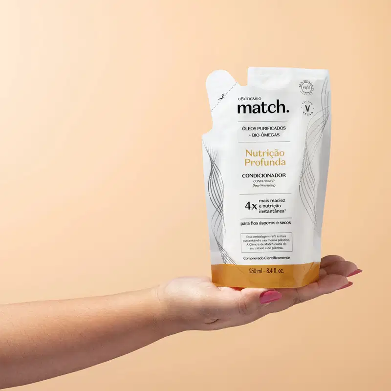 Imagem de uma mão segurando uma sachê refil do condicionador Match Nutrição Profunda. A embalagem é branca com detalhes em laranja  e letras pretas. O fundo possui tonalidades de rosa.