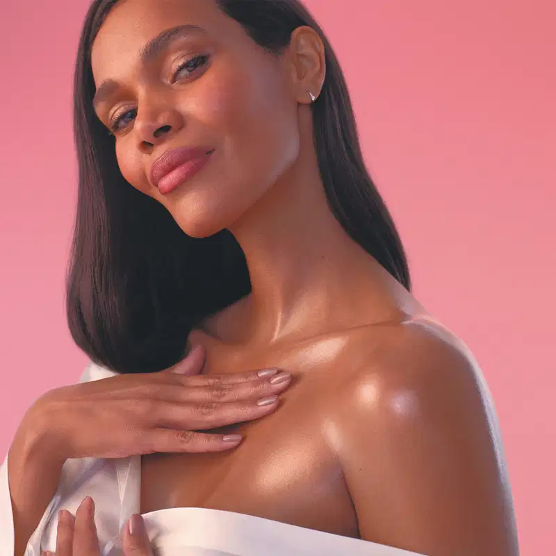 Mulher de pele negra com expressão neutra, cabelos longos, lisos e pretos, usando uma camisa branca desabotoada mostrando um dos ombros, colo e pescoço. Sua pele está luminosa e uma das mãos toca o ombro, em um fundo rosa.