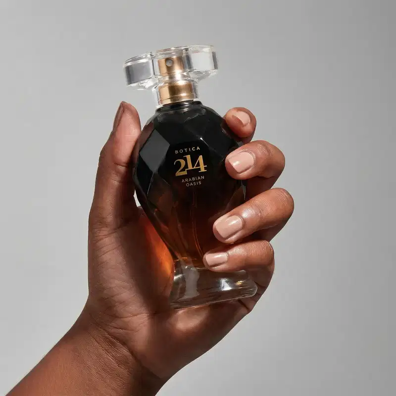 Frasco de item de perfumaria da marca Botica 214 Arabian Oasis sendo segurado firmemente por mãos femininas sobre fundo cinza, evidenciando nome em dourado gravado no vidro escuro.