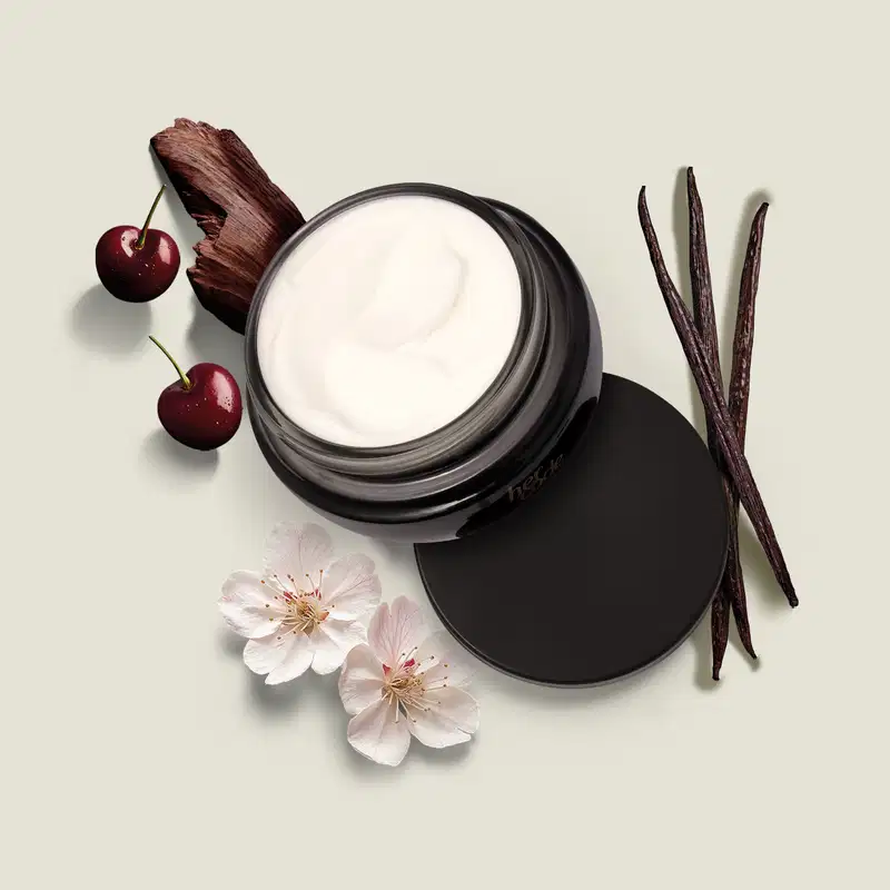 Ao redor do pote preto com creme branco contendo creme hidratante corporal da marca Her Code, há alguns elementos decorativos, como cereja, flores e uma vagem de baunilha. A imagem tem um fundo claro e neutro, destacando o produto central.