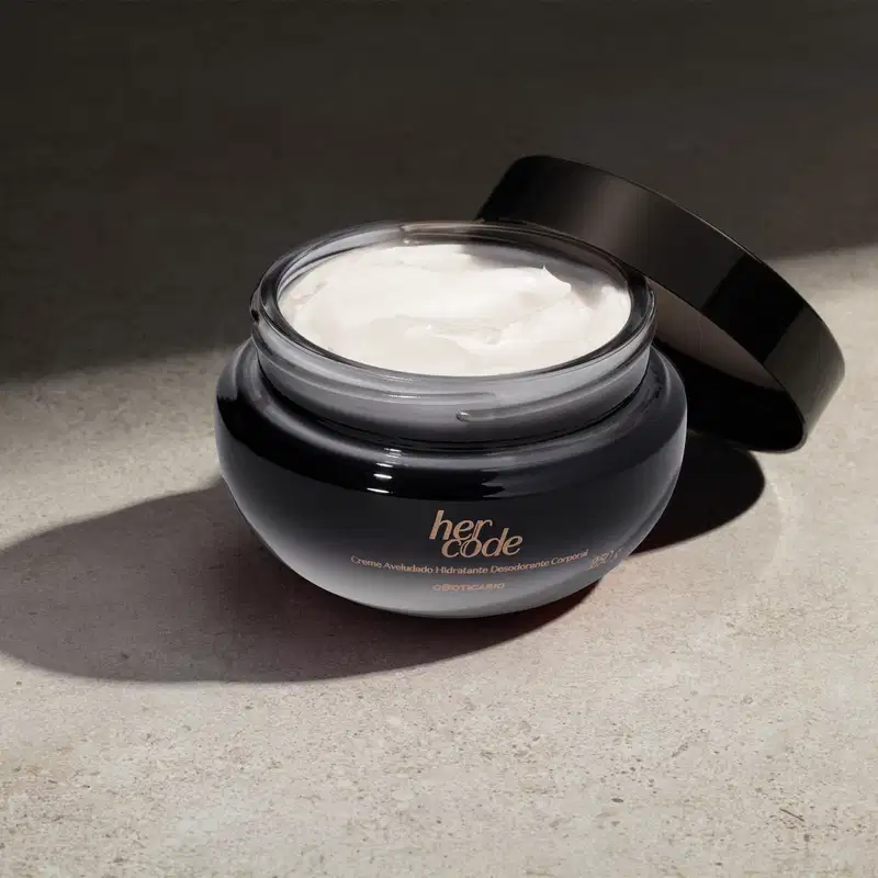 O produto é um creme hidratante desodorante da linha Her Code, em uma embalagem de vidro com tampa preta. O creme em si tem uma aparência cremosa e branca, preenchendo o recipiente de vidro. Está em fundo escuro com iluminação na superficie.