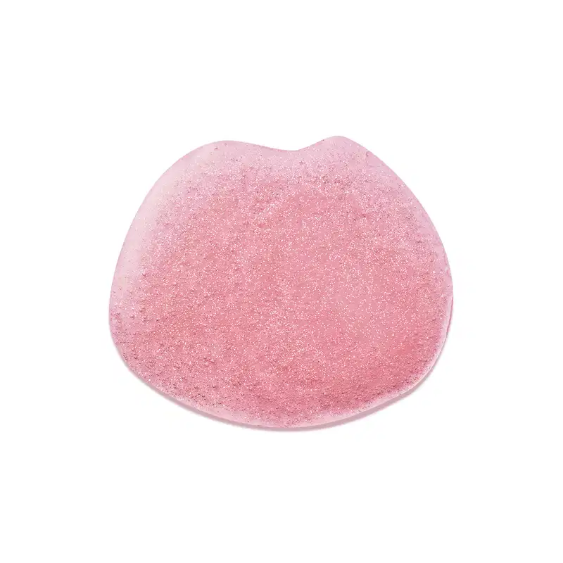 Objeto de perfumaria com textura granulada e brilhante em tom de rosa. Superfície irregular e aspecto cristalino, sugerindo uma composição sólida e possivelmente relacionada a produtos de beleza.