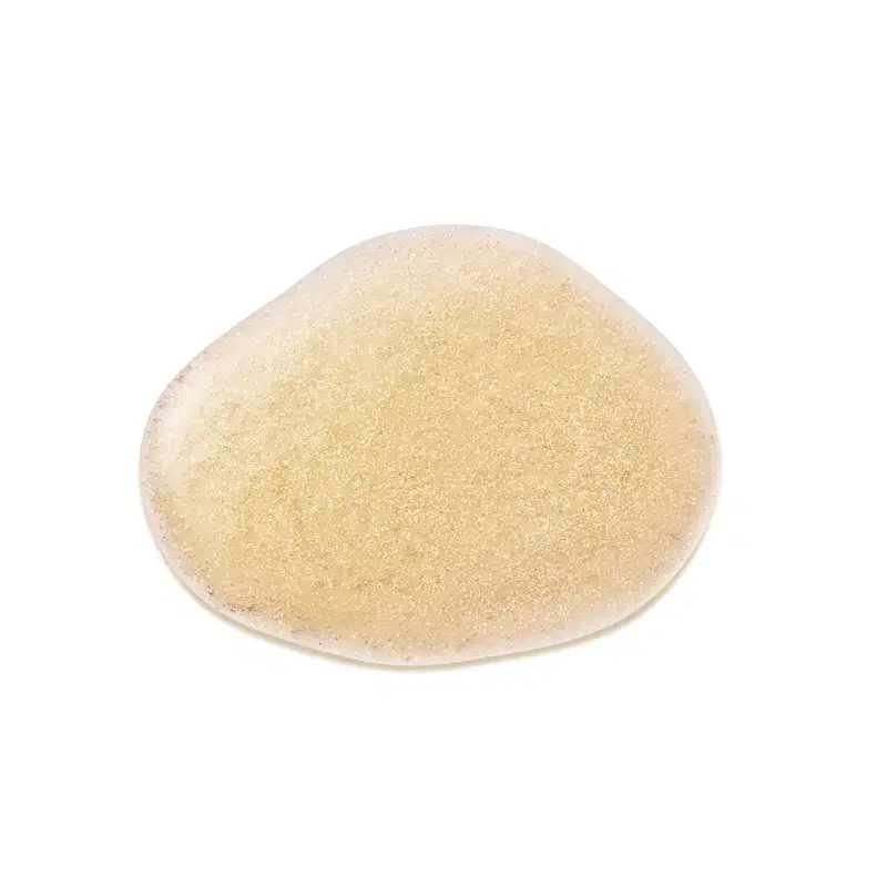 Objeto de textura granulada, com coloração amarelo-clara. Aparenta ser um produto de beleza, possivelmente um item de perfumaria. Sua superfície apresenta uma consistência sólida e brilhante, sugerindo uma textura arenosa ou cristalina.