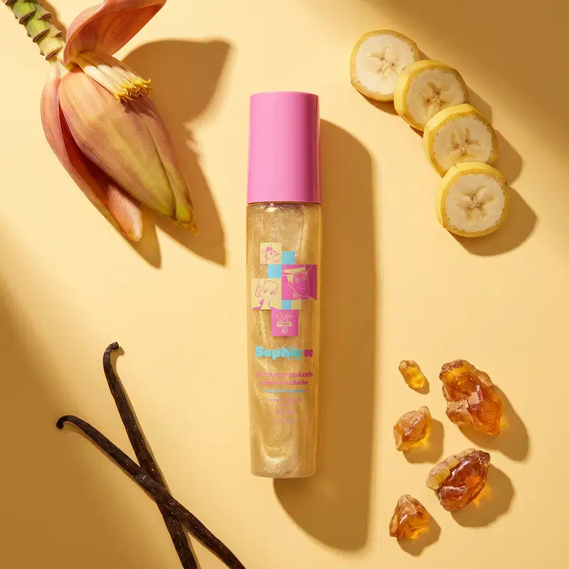 Ambiente de beleza com flores, frutas e cristais âmbar. Embalagem rosa e dourada de um item de perfumaria da marca Sophie, com ilustrações e texto "Toy Story 5". Fundo amarelo ilumina os elementos naturais.