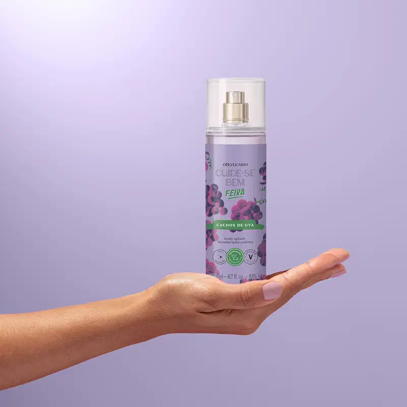 Imagem de um frasco de body splash da marca Cuide-se bem Feira Cachos de Uva com rótulo roxo e ilustração de uvas roxas, seguro por mão em fundo roxo claro.