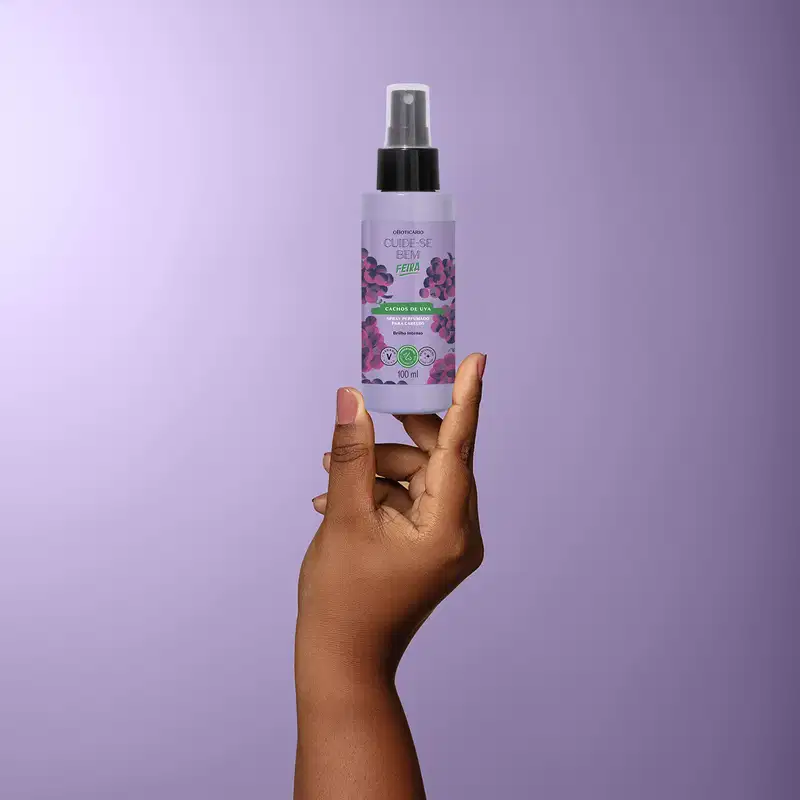 Frasco roxo de spray perfumado para cabelos com rótulo roxo e imagem de uvas, em mão contra fundo roxo claro.