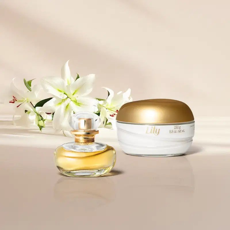 Imagem de um frasco dourado de Eau de Parfum Lily e um pote branco com tampa dourada de creme corporal da mesma marca em fundo e superfície claras e lírios brancos ao lado.